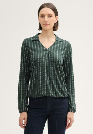 Grüne Bluse mit langen Ärmeln und V-Ausschnitt, mit vertikalen weißen Streifen und einer weichen Textur. Kombiniert mit dunklen Jeans.