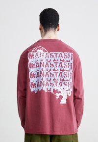 Burgunderfarget langermet skjorte med et stort hvitt grafisk design på baksiden som viser gjentatt tekst og et illustrativt design av et tre.