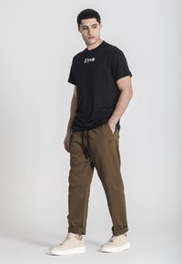 Černé tričko s krátkým rukávem se logem, v kombinaci s hnědými kalhotami se stahovacími šňůrkami v pase. Model má na sobě béžové tenisky. Jednoduché pozadí.