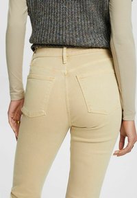 Vaqueros de talle alto en color beige con un corte ajustado, que presentan dos bolsillos traseros y detalles de costura sutiles. Se llevan con una blusa de manga larga de color claro.