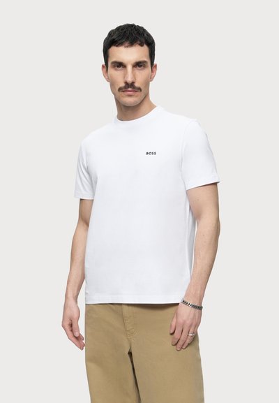 Homme aux cheveux courts et foncés et à la moustache, portant un T-shirt BOSS blanc et un pantalon beige, debout devant un fond clair et uni.
