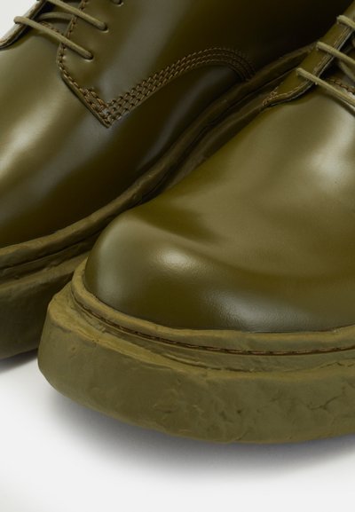 Zapatos de cuero verde con un acabado suave, punta redonda y suela de goma texturada gruesa. Presentan costuras marrones a lo largo de la parte superior.
