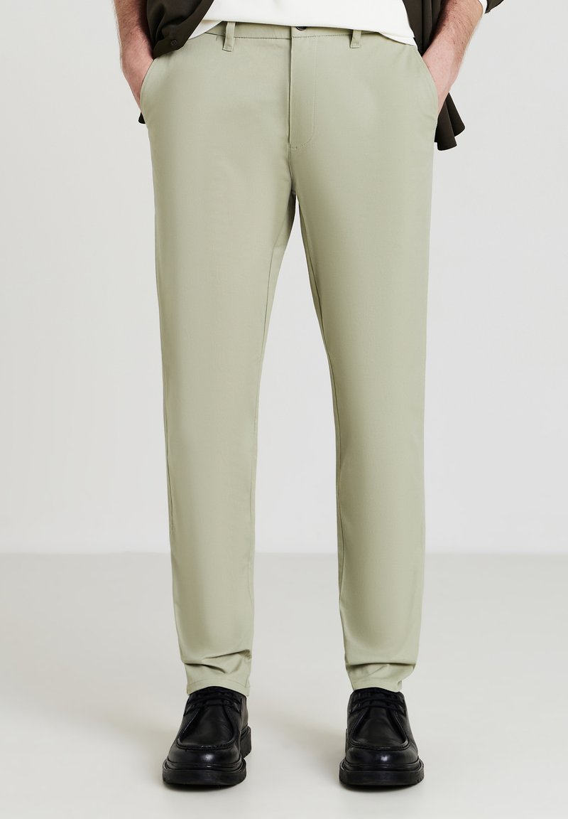 Light green tailored pants with a smooth texture, front pockets, and a fitted design, paired with black shoes.