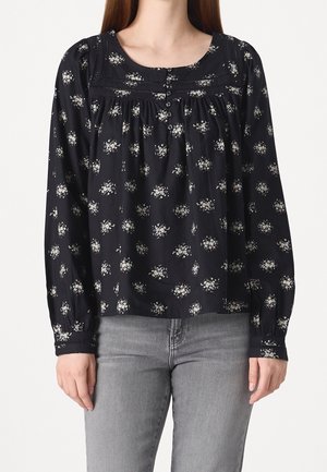 Kvinde iført en sort langærmet bluse med små hvide blomsterprint og knapper, kombineret med lysegrå jeans.