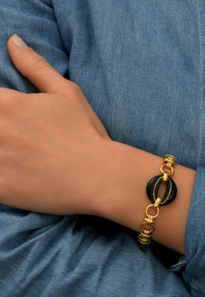 Bracelet en chaîne dorée avec des accents circulaires noirs, présentant des maillons entrelacés et des détails texturés, porté sur une main contre un tissu en denim.