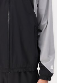 Black and gray zip-up jacket with elastic waistband and cuffs; smooth, lightweight fabric; features a side pocket and subtle logo detail.