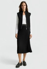 Gilet senza maniche nero a coste sopra una lunga gonna nera plissettata, abbinati a scarpe platform nere. Texture liscia e design moderno.