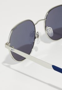 Στρογγυλά γυαλιά ηλίου με σκούρους φακούς, ασημένιο μεταλλικό σκελετό και μπλε λεπτομέρειες στους βραχίονες. Διαθέτουν επένδυση στη μύτη για περισσότερη άνεση.