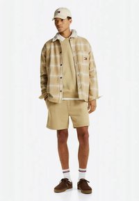 Man wearing beige plaid jacket, tan sweatshirt, shorts, white cap, white socks, and brown sneakers standing against white background.