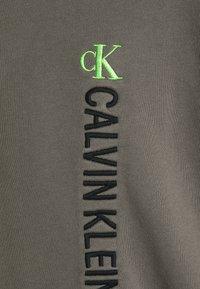 Tejido gris con bordado vertical negro que presenta "CALVIN KLEIN" y un pequeño logotipo verde "CK". Textura suave, diseño sencillo.