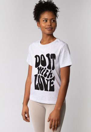 Vrouw met krullend haar die een wit t-shirt draagt met grote zwarte tekst 'DO IT with LOVE' en lichtgekleurde geribbelde broek, kijkt opzij.