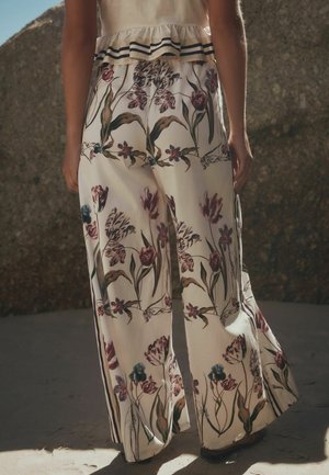 Person, die cremefarbene, weit geschnittene Hosen mit detailliertem lila-grünem Blumenmuster trägt, steht draußen auf sandigem Boden in der Nähe einer felsigen Oberfläche.