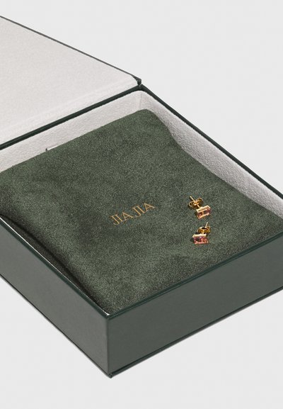 Zelta toņa auskari ar taisnstūra rozā akmeņiem, iesaiņoti zaļā samta maisiņā tumši zaļā kastītē, uz kuras ir iepresēts "JIA JIA."