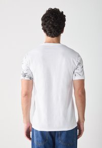 White short-sleeve t-shirt with floral patterned sleeves, featuring a round neckline and a smooth texture, worn with blue jeans.