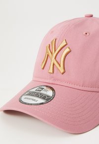 Rozā kokvilnas cepure ar regulējamu siksniņu, uz kuras priekšpuses ir zeltā izšūta "NY" logo un apļa brenda uzlīme uz malas.