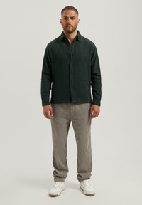 Man die staat te poseren in een donkergroene, gestructureerde overhemd met knopen, bruin geruite broek en witte sneakers tegen een effen achtergrond.