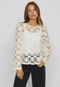 Haut en dentelle florale blanche avec un design transparent, des manches longues et un col rond. Superposé sur une couche intérieure blanche unie.