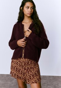 Cárdigan de punto burdeos oscuro con cierre de cremallera al frente, mangas largas y textura acanalada, llevado sobre un vestido marrón con estampado y dobladillo con volantes.