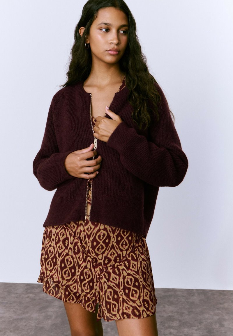 Cárdigan de punto burdeos oscuro con cierre de cremallera al frente, mangas largas y textura acanalada, llevado sobre un vestido marrón con estampado y dobladillo con volantes.