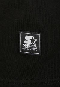 Tissu noir avec une étiquette rectangulaire cousue comportant un grand "S" avec une étoile et le texte "STARTER BLACK LABEL" en blanc.