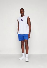 White sleeveless top with blue 'LA' logo, paired with blue shorts. Model wears white socks and sneakers. Simple athletic design.
