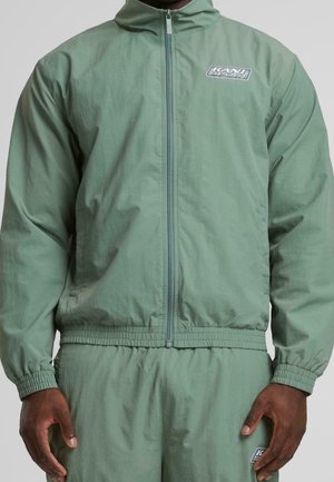 Hombre con chaqueta con cremallera verde salvia y pantalones a juego con puños elásticos, con pequeños logotipos "KANI SPORT" en el pecho y el muslo.