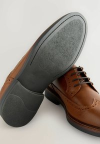 Zapato brogue de cuero marrón con un upper texturizado, detalles perforados y una suela de goma negra con un acabado suave y forma redondeada.
