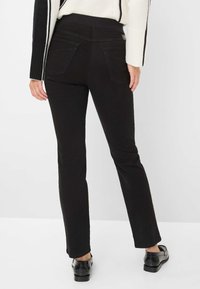 Schwarze, figurbetonte Jeans mit glatter Textur, mittelhohem Bund und Details an den Gesäßtaschen. Kombiniert mit schwarzen Schuhen vor neutralem Hintergrund.