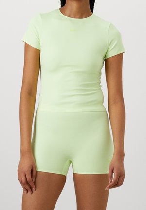 Femme portant un haut ajusté à manches courtes vert clair et un short taille haute assorti, devant un fond gris uni.