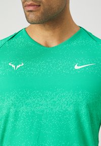 Zelena sportska majica s teksturiranim uzorkom, okruglim izrezom i dva logotipa: bijeli Nike swoosh i silueta bika.