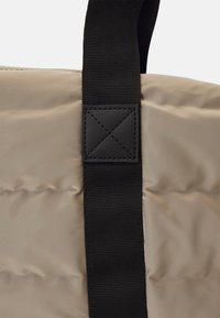 Sac rembourré beige avec une bandoulière noire. Présente une surface texturée et matelassée ainsi qu'un accent en cuir noir sur la bandoulière avec des détails cousus.