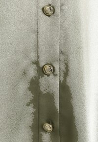 Olivgrön satinblus med metallisk glans, med gyllene knappar och ett vattenliknande mönster över tyget.