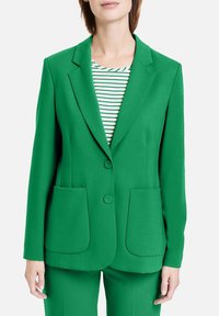 Blazer verde con un diseño estructurado, dos bolsillos frontales, solapas con muescas y pantalones verdes a juego. Debajo hay una camiseta a rayas.