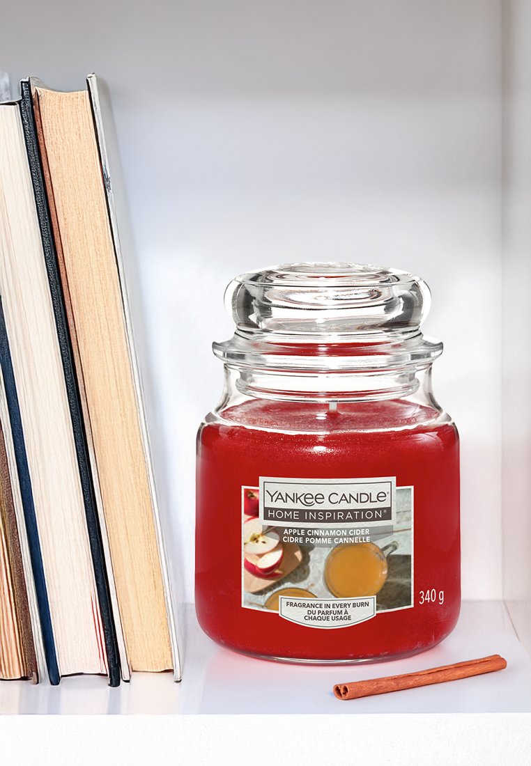 Candelina in vaso di vetro rosso con coperchio trasparente, etichettata "Sidro di Mela e Cannella." Posizionata accanto a dei libri, include un bastoncino di cannella come accentuazione.