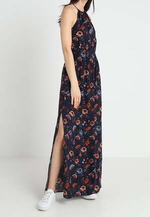 Maxikleid mit floralem Muster in Blau, Rot und Grün auf dunklem Hintergrund. Mit Neckholder, elastischer Taille und Seitenschlitz. Gekleidet mit weißen Turnschuhen.