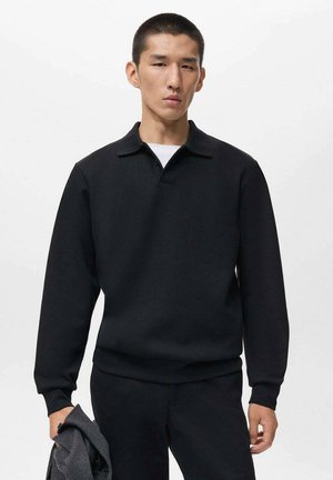 Asiatischer Mann, der ein schwarzes Langarmpolo-Shirt und schwarze Hosen trägt, während er eine gefaltete graue Jacke vor einem schlichten weißen Hintergrund hält.