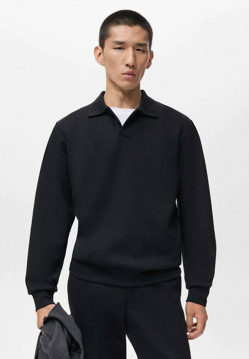 Hombre asiático con una camisa polo negra de manga larga, pantalones negros, sosteniendo una chaqueta gris doblada contra un fondo blanco liso.