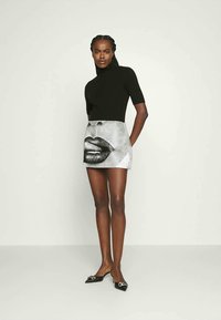 Top negro ajustado con cuello alto, combinado con una minifalda metálica que presenta un gráfico monocromo de labios. La modelo lleva unos zapatos planos negros con detalle de lazo.