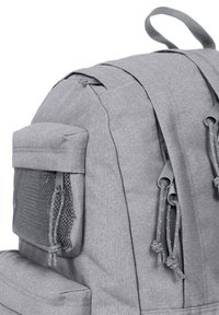 Mochila de tela gris con múltiples compartimentos con cremallera y un bolsillo lateral de malla, que cuenta con cordones en bucle en las cremalleras y un asa superior para transportar.