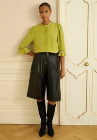 Limonkowa bluzka z bufkami i plisami, zestawiona z czarnymi, wysokimi spodniami wystającymi z materiału skórzanego i czarnymi, wysokimi, spiczastymi botkami.