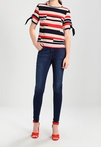 Top de mangas cortas con rayas horizontales en rojo, azul marino y beige, con detalles de lazo en las mangas, acompañado de jeans skinny oscuros y tacones rojos.