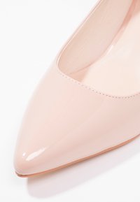 Chaussure féminine brillante rose pâle à bout pointu avec coutures visibles sur la tige et bord de semelle marron clair sur fond blanc.