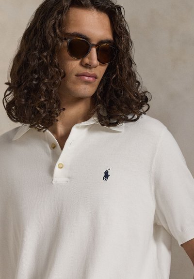 Άνδρας με σγουρά μαλλιά που φοράει γυαλιά ηλίου με σχέδιο χελώνας και λευκό πόλο πουκάμισο με μικρό σκούρο μπλε κεντημένο λογότυπο στο στήθος.