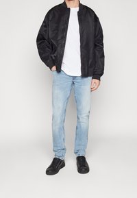 Chaqueta bomber negra, material ligero, puños acanalados, camiseta blanca debajo, vaqueros azul claro, zapatillas negras, estilo casual.