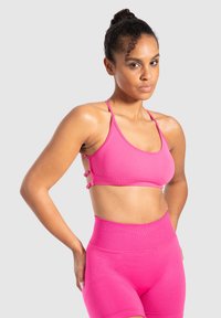 Vrouw met krullend haar in een hoge bun, gekleed in een felroze sportbeha en hoogstelige shorts, staand tegen een lichtgrijze achtergrond.