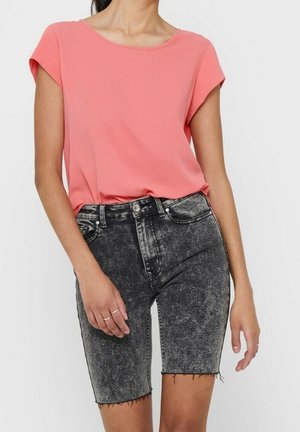 Vrouw draagt een koraalkleurig blouse met korte mouwen, ingestopt in een hoge taille, zwarte denim shorts met een zuur-was effect en rafelige zoom.