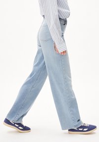 Person går iført lys blå bredbenede jeans, blå sneakers med hvide detaljer, og en langærmet skjorte med blå og hvide striber.