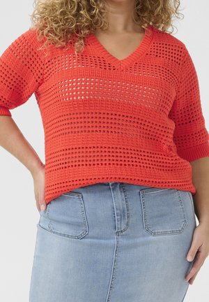 Červený pletený svetr s výstřihem do V a krátkými rukávy, zdobený vzorovanou otevřenou vazbou. Kombinovaný se světle modrou džínovou sukní.