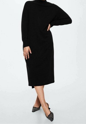 Kobieta stoi z jedną ręką na biodrze, ubrana w długą czarną sukienkę i czarne sandały typu slingback z białym wzorem.