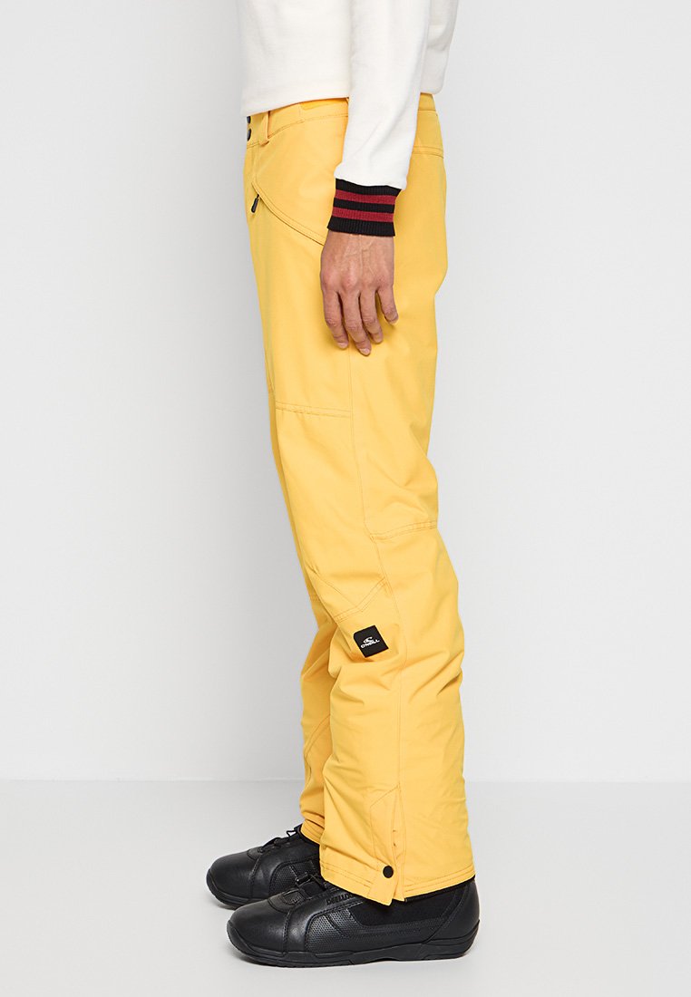 Gelbe Snowboardhose mit schmalem Schnitt, ausgestattet mit einer seitlichen Reißverschlusstasche und einem Logo-Patch. Kombiniert mit schwarzen Sportschuhen.